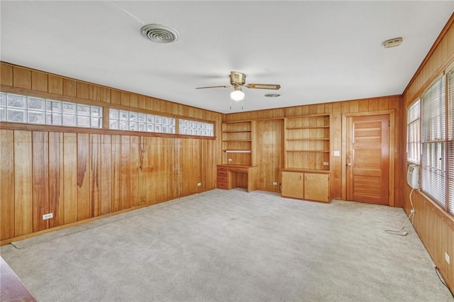 empty room with built in shelves, wood walls, light colored carpet, and ceiling fan