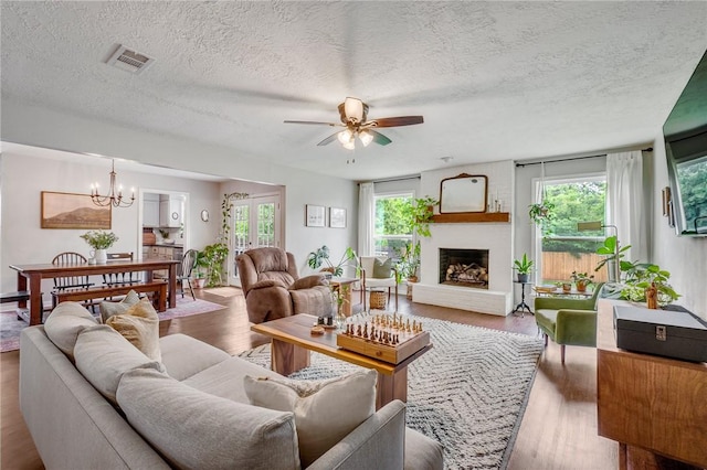 living area featuring a textured ceiling, visible vents, a brick fireplace, wood finished floors, and ceiling fan with notable chandelier