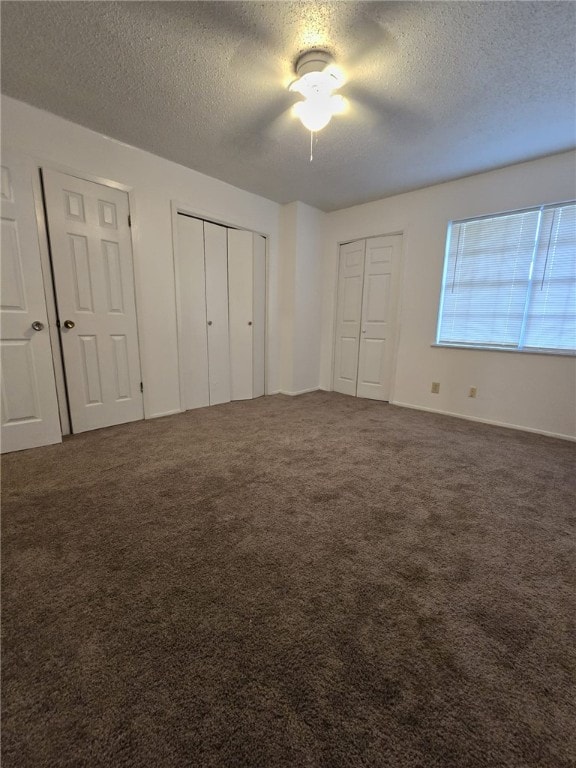 unfurnished bedroom featuring a textured ceiling, ceiling fan, dark carpet, and two closets