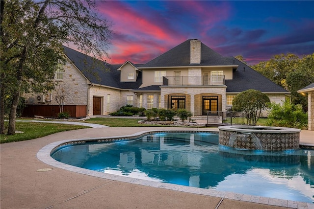 back house at dusk featuring a lawn, a swimming pool with hot tub, and a balcony