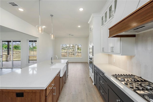 kitchen with appliances with stainless steel finishes, an island with sink, white cabinets, decorative light fixtures, and custom exhaust hood