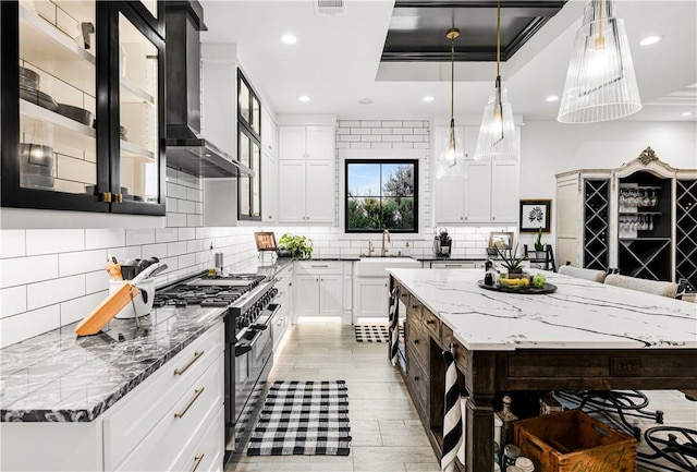 kitchen with white cabinets, backsplash, light stone countertops, wall chimney exhaust hood, and gas range