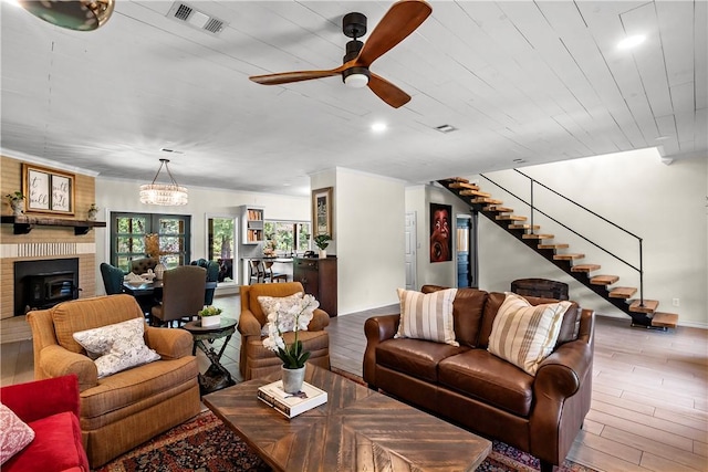 living room with wood finished floors, ceiling fan with notable chandelier, a brick fireplace, visible vents, and stairs