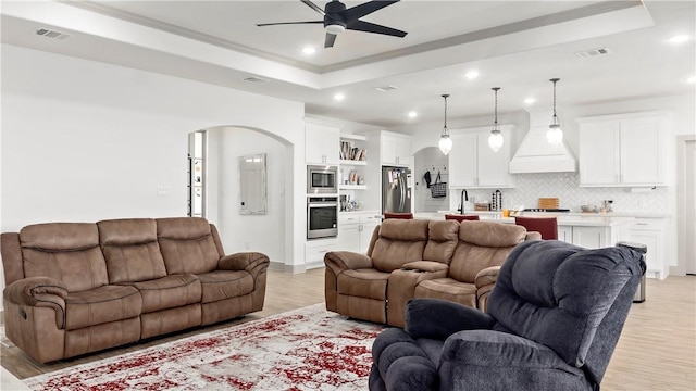 living room with a tray ceiling, ceiling fan, sink, and light wood-type flooring