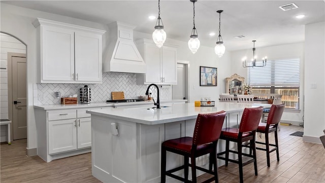 kitchen with premium range hood, white cabinetry, hanging light fixtures, and a center island with sink