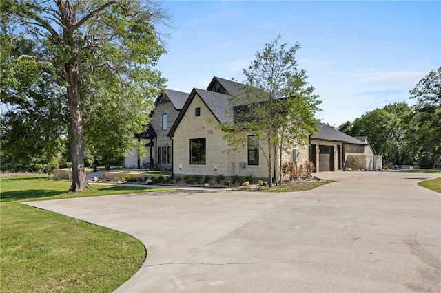 french country inspired facade with roof with shingles, driveway, stone siding, an attached garage, and a front lawn