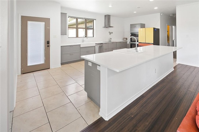 kitchen with gray cabinetry, wall chimney range hood, sink, an island with sink, and appliances with stainless steel finishes