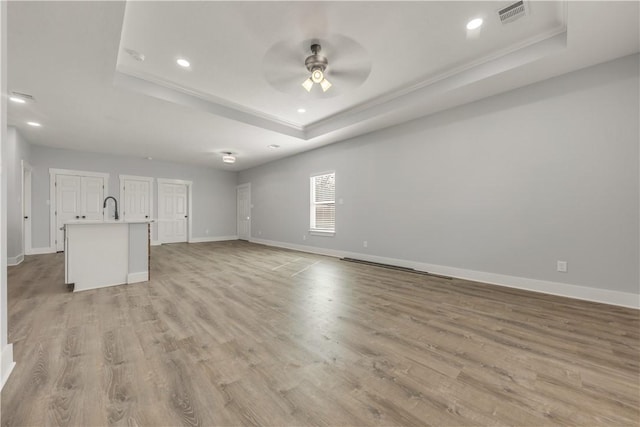 unfurnished living room featuring sink, ceiling fan, a tray ceiling, crown molding, and light wood-type flooring