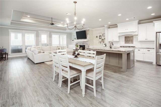 dining room featuring ceiling fan with notable chandelier, a tray ceiling, sink, a fireplace, and light hardwood / wood-style floors