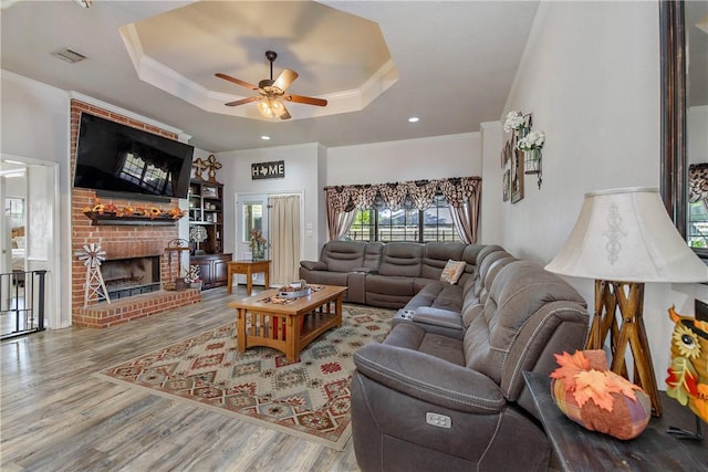 living room featuring hardwood / wood-style floors, a brick fireplace, ceiling fan, ornamental molding, and a tray ceiling