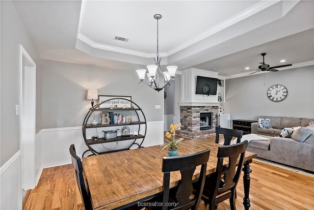 dining space with light wood-type flooring, a brick fireplace, ornamental molding, ceiling fan with notable chandelier, and a raised ceiling