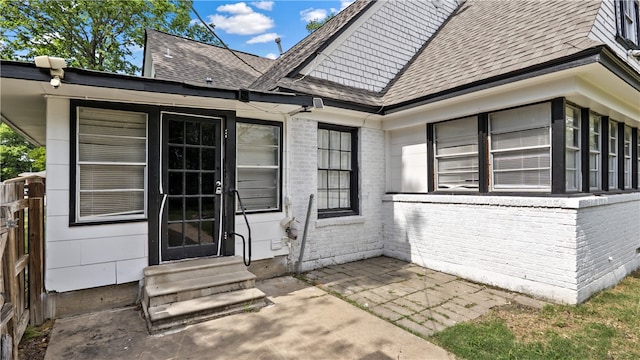 view of exterior entry with brick siding, a shingled roof, a patio, and fence
