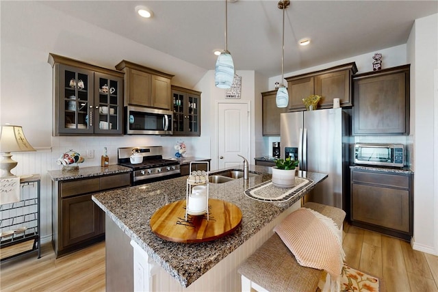 kitchen featuring light hardwood / wood-style floors, stainless steel appliances, a kitchen island with sink, dark stone countertops, and hanging light fixtures