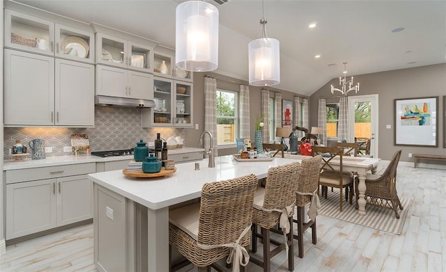 kitchen featuring a center island with sink, glass insert cabinets, light countertops, under cabinet range hood, and pendant lighting