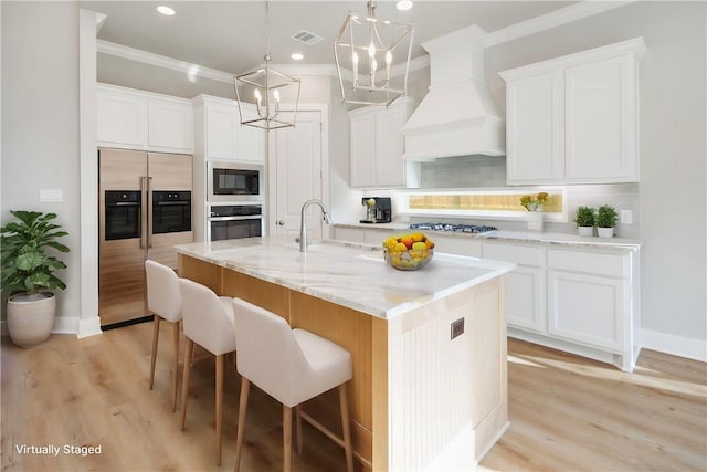 kitchen featuring premium range hood, crown molding, built in appliances, a center island with sink, and white cabinetry