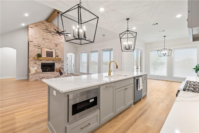 kitchen with a brick fireplace, stainless steel appliances, pendant lighting, gray cabinets, and an island with sink