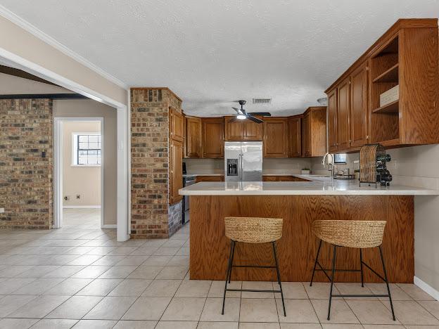 kitchen with ceiling fan, stainless steel refrigerator with ice dispenser, kitchen peninsula, crown molding, and light tile patterned flooring