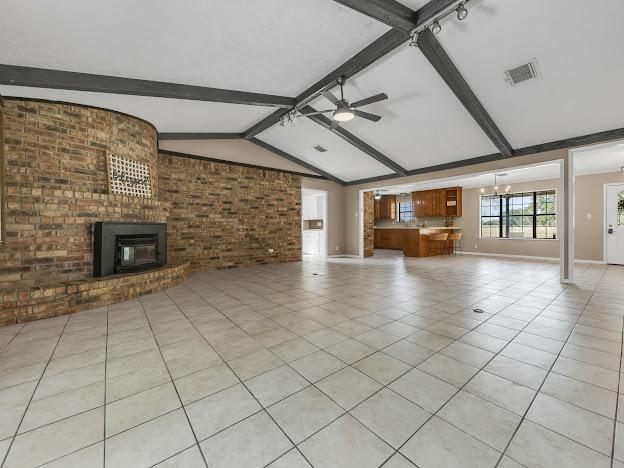 unfurnished living room featuring brick wall, ceiling fan with notable chandelier, light tile patterned floors, lofted ceiling with beams, and a fireplace