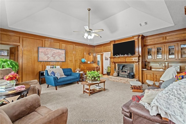 living room with carpet flooring, a raised ceiling, ceiling fan, a wood stove, and wood walls