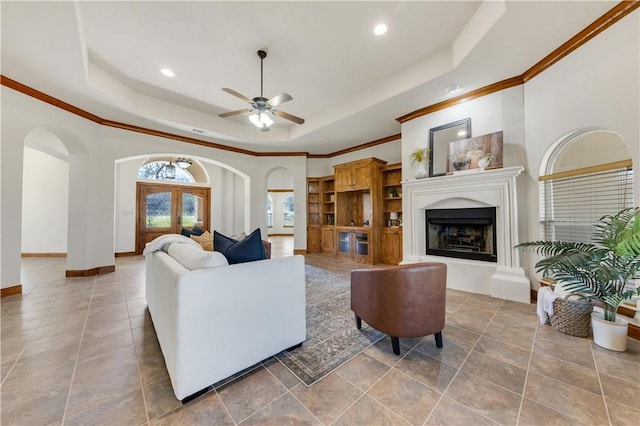 tiled living room with a fireplace with raised hearth, crown molding, baseboards, arched walkways, and a raised ceiling