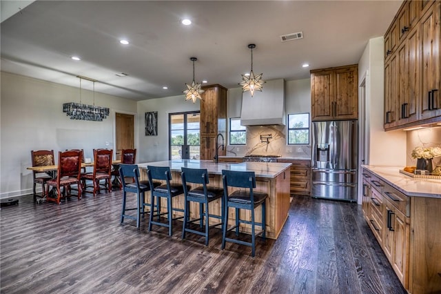 kitchen featuring a large island with sink, custom exhaust hood, decorative light fixtures, dark wood-type flooring, and stainless steel fridge with ice dispenser