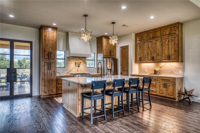 kitchen featuring pendant lighting, custom exhaust hood, dark wood-type flooring, stainless steel fridge, and a kitchen island with sink