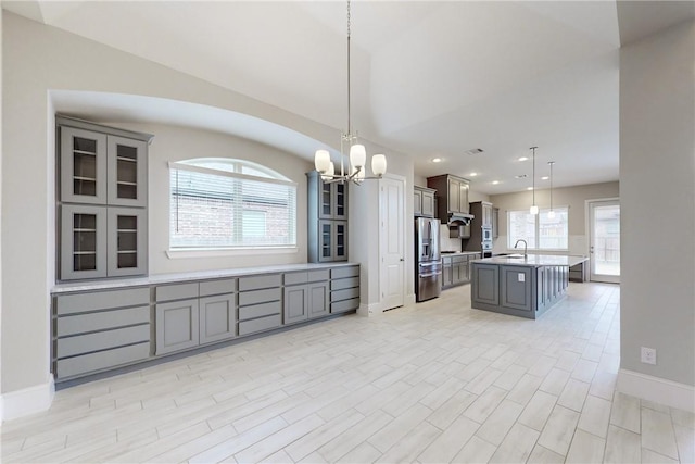 kitchen featuring gray cabinetry, stainless steel refrigerator with ice dispenser, sink, decorative light fixtures, and an island with sink