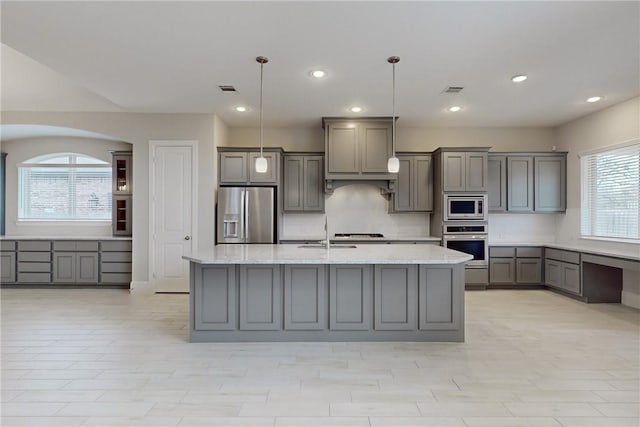 kitchen featuring appliances with stainless steel finishes, hanging light fixtures, a kitchen island with sink, light stone counters, and gray cabinets