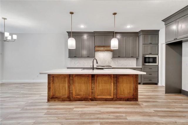 kitchen with gray cabinetry, a kitchen island with sink, stainless steel microwave, sink, and hanging light fixtures