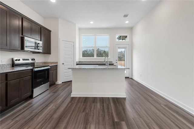 kitchen with stainless steel appliances, an island with sink, light stone countertops, and dark brown cabinetry