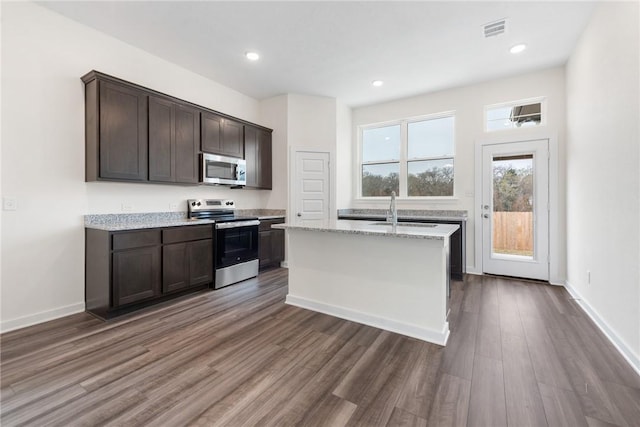 kitchen with light stone counters, dark brown cabinets, stainless steel appliances, and an island with sink