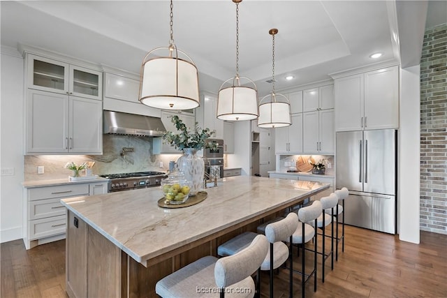 kitchen with wall chimney range hood, premium appliances, an island with sink, decorative light fixtures, and white cabinets