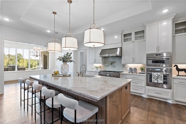 kitchen with sink, wall chimney exhaust hood, an island with sink, white cabinets, and appliances with stainless steel finishes