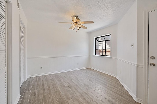 unfurnished bedroom with a textured ceiling, light wood-style flooring, a wainscoted wall, visible vents, and a ceiling fan