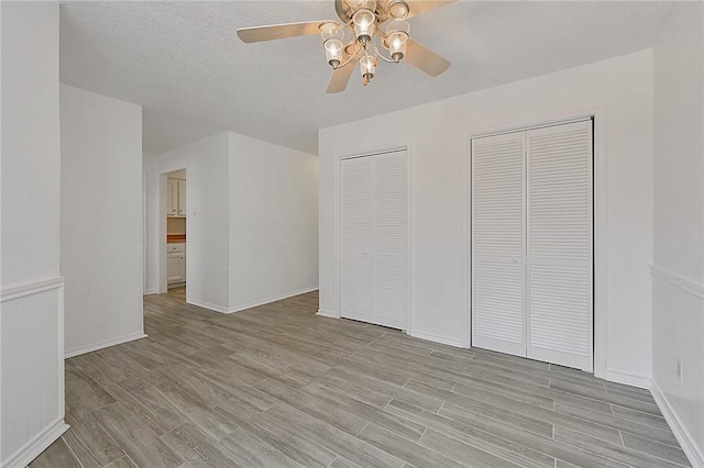 unfurnished bedroom with a textured ceiling, ceiling fan, light wood-style flooring, baseboards, and multiple closets