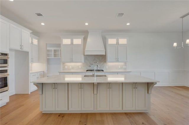 kitchen featuring premium range hood, white cabinetry, stainless steel oven, a center island with sink, and light hardwood / wood-style flooring