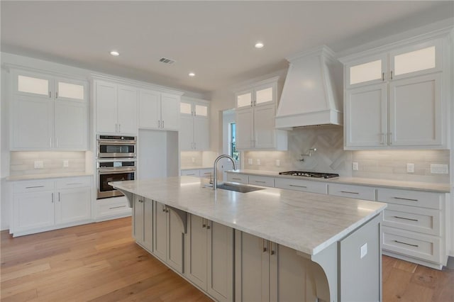 kitchen with premium range hood, sink, a center island with sink, stainless steel appliances, and white cabinets