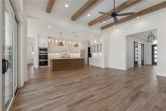 unfurnished living room featuring wood-type flooring, sink, ceiling fan, a barn door, and beam ceiling