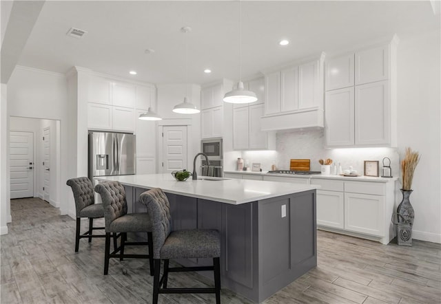 kitchen with appliances with stainless steel finishes, a center island with sink, light hardwood / wood-style flooring, white cabinets, and hanging light fixtures