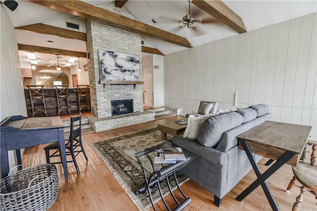 living room featuring wood walls, lofted ceiling with beams, a brick fireplace, ceiling fan, and light hardwood / wood-style floors