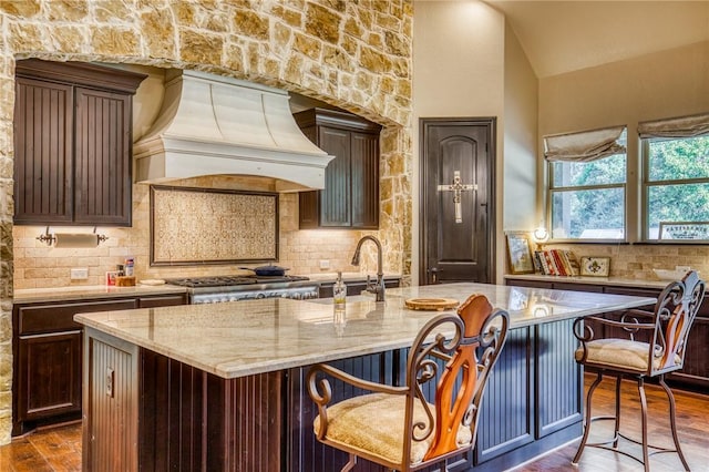 kitchen featuring tasteful backsplash, a center island with sink, dark wood-style floors, light stone countertops, and premium range hood