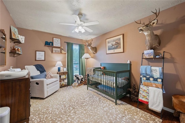 bedroom with a crib, a textured ceiling, a ceiling fan, baseboards, and wood finished floors