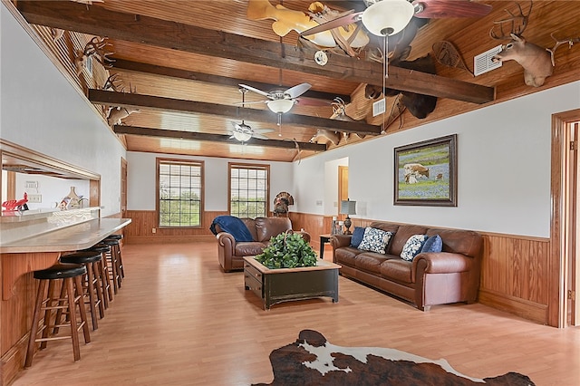 living room featuring light wood-style flooring, wooden walls, a wainscoted wall, a ceiling fan, and beamed ceiling