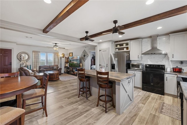 kitchen with open floor plan, black / electric stove, wall chimney range hood, white cabinetry, and stainless steel refrigerator with ice dispenser