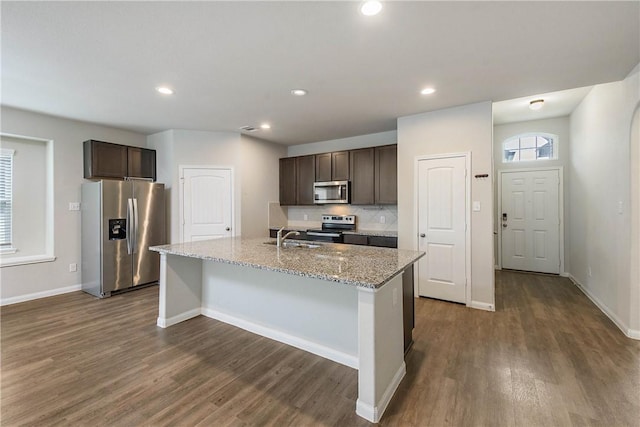 4706 Coyotillo Way, Bryan, TX 77807 - kitchen with appliances with stainless steel finishes, dark brown cabinetry, dark wood-type flooring, a kitchen island with sink, and light stone counters