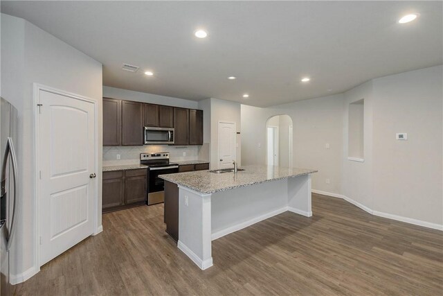 4706 Coyotillo Way, Bryan, TX 77807 - kitchen featuring appliances with stainless steel finishes, arched walkways, dark wood-type flooring, an island with sink, and dark brown cabinets
