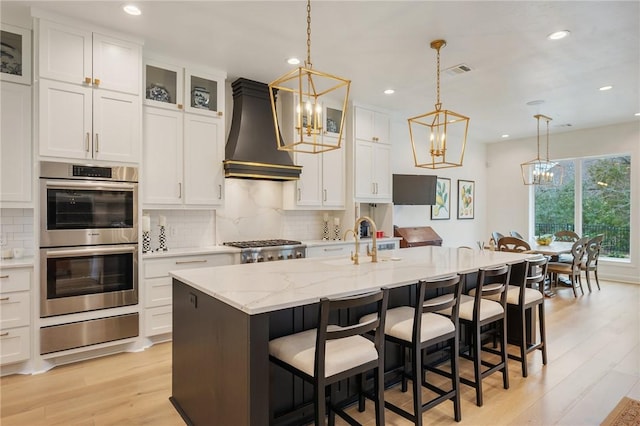 kitchen with double oven, decorative light fixtures, premium range hood, a center island with sink, and glass insert cabinets