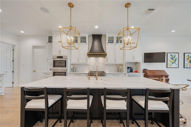 kitchen with custom range hood, visible vents, glass insert cabinets, a large island with sink, and a kitchen breakfast bar