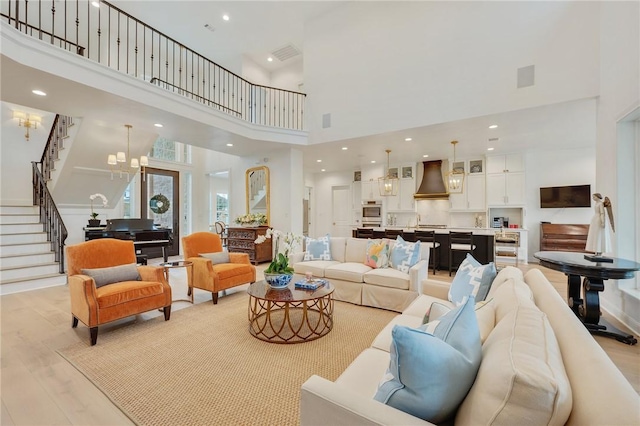 living room featuring a towering ceiling, light wood-style flooring, an inviting chandelier, stairs, and recessed lighting