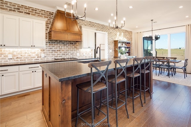 kitchen with light wood-type flooring, custom range hood, a kitchen island with sink, white cabinetry, and hanging light fixtures